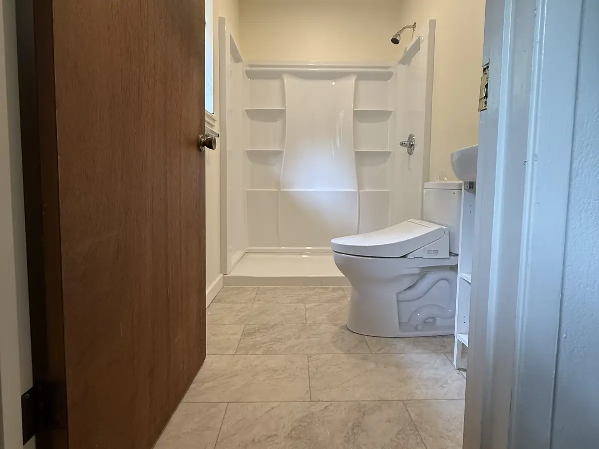 A bathroom with a white shower, a toilet and a tile floor, seen through the open door