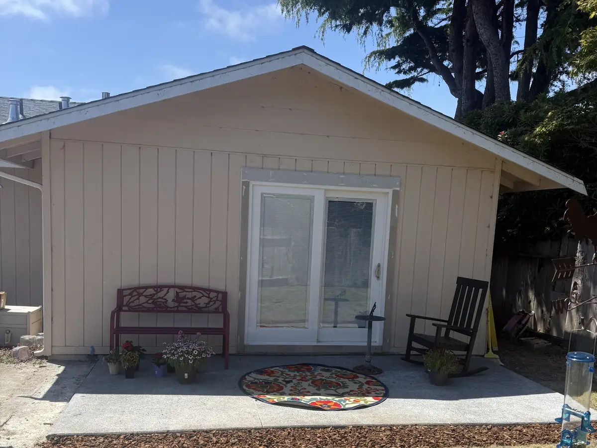 A small building with sliding glass doors, a bench and a rocking chair on a concrete patio